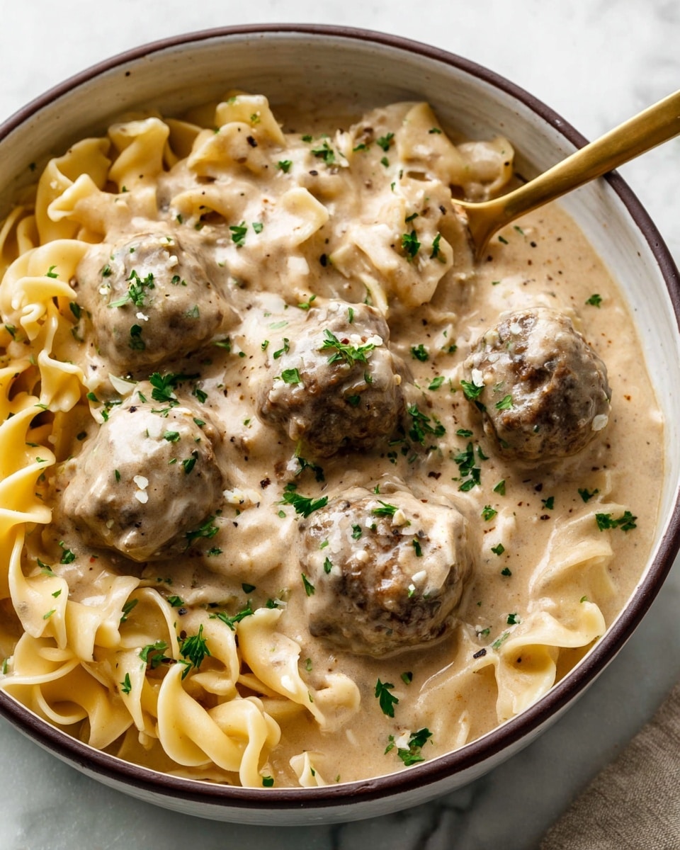 A close-up image of a bowl filled with creamy stroganoff. The dish has two main layers: the first layer is the cooked egg noodles that are light yellow, twisted, and soft, sitting at the bottom and edges of the bowl. The second layer is a thick, creamy beige sauce covering the noodles and containing several browned meatballs scattered around. The sauce has a smooth texture with visible specks of black pepper and finely chopped green parsley sprinkled on top, adding color contrast. A gold spoon is scooping one meatball from the bowl. The bowl is white with a dark brown rim and sits on a white marbled surface. photo taken with an iphone --ar 4:5 --v 7