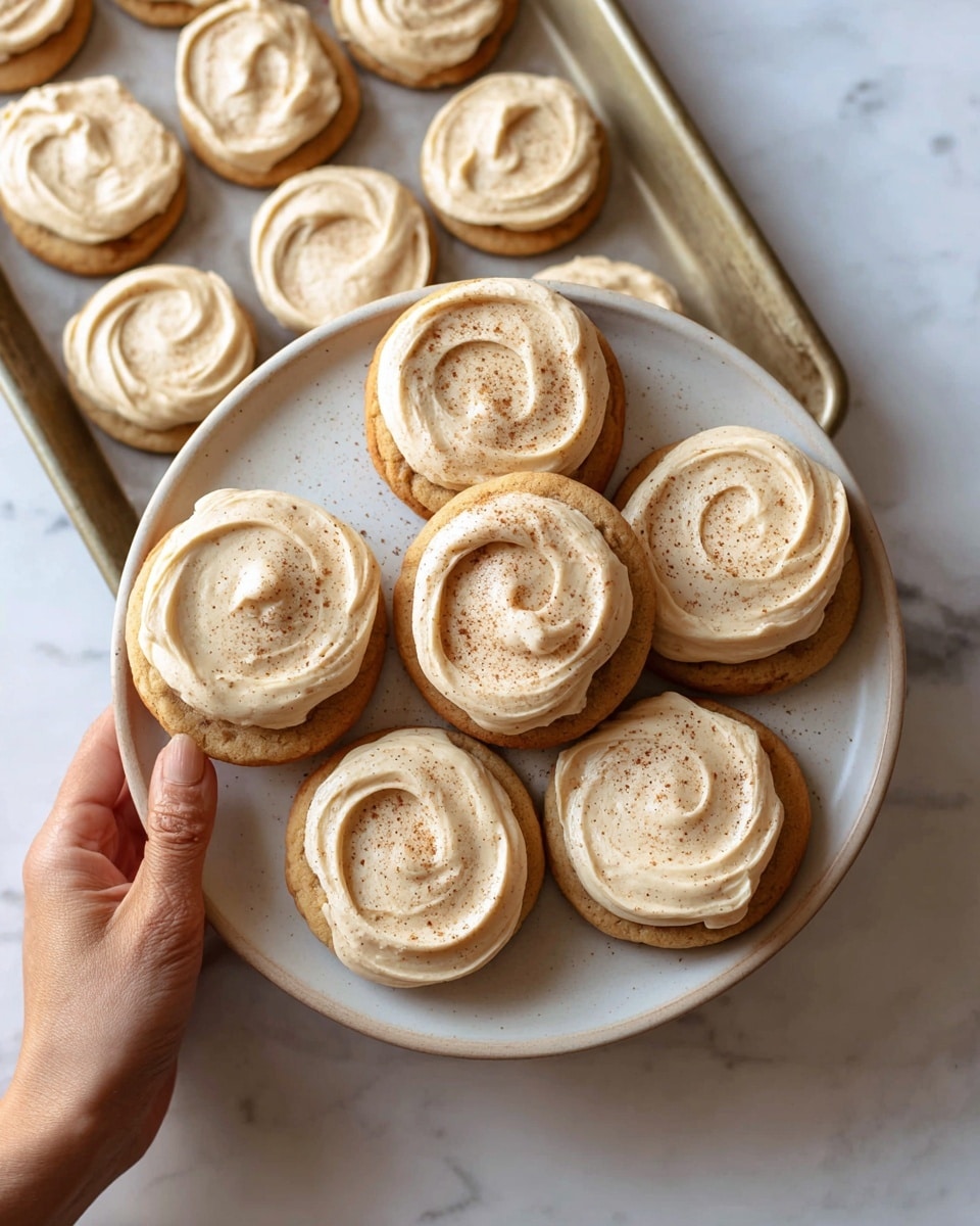 A close-up view of several round cookies arranged on a white plate, each cookie topped with one thick layer of creamy light brown frosting that has visible swirls and a soft texture, lightly sprinkled with fine darker brown powder, set on a white marbled surface with a folded rust-colored cloth partially visible in the background, photo taken with an iphone --ar 4:5 --v 7