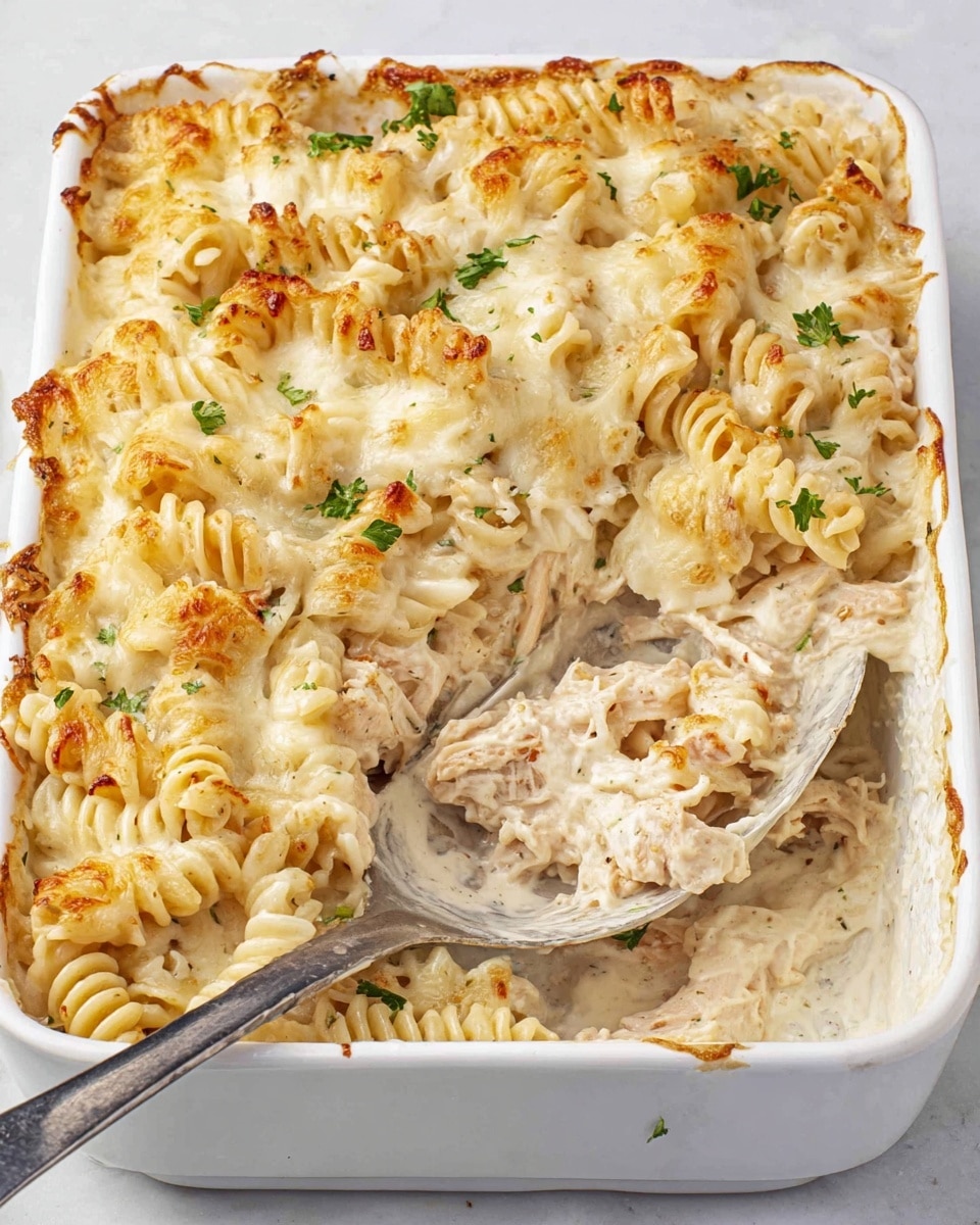 A white rectangular baking dish holds creamy baked pasta made with short spiral noodles coated in a thick, light beige cream sauce. The top layer shows melted cheese browned slightly in golden spots, giving a bubbly and soft texture with small browned edges. Small green parsley leaves are scattered lightly on top, adding color contrast. A large silver serving spoon rests inside the dish, partially scooping out some pasta, revealing the creamy layers beneath, with visible bits of cooked chicken mixed throughout the spirals. The dish sits on a white marbled surface. photo taken with an iphone --ar 4:5 --v 7