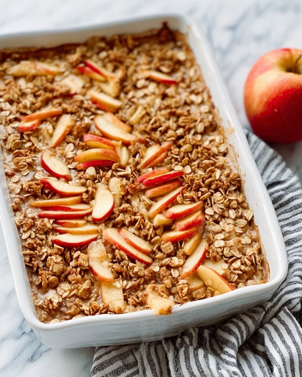A white rectangular dish holds a baked apple dessert with many layers. The top layer is made up of small apple slices with red skin, lightly browned and mixed with crunchy oat topping that is golden-brown. You can see pieces of nuts or granola sprinkled on top, giving it a rough texture. The dessert looks warm and soft underneath the crisp oat layer. To the side of the dish, there is a fresh red apple. The background is a white marbled surface with a striped cloth under the dish. photo taken with an iphone --ar 4:5 --v 7