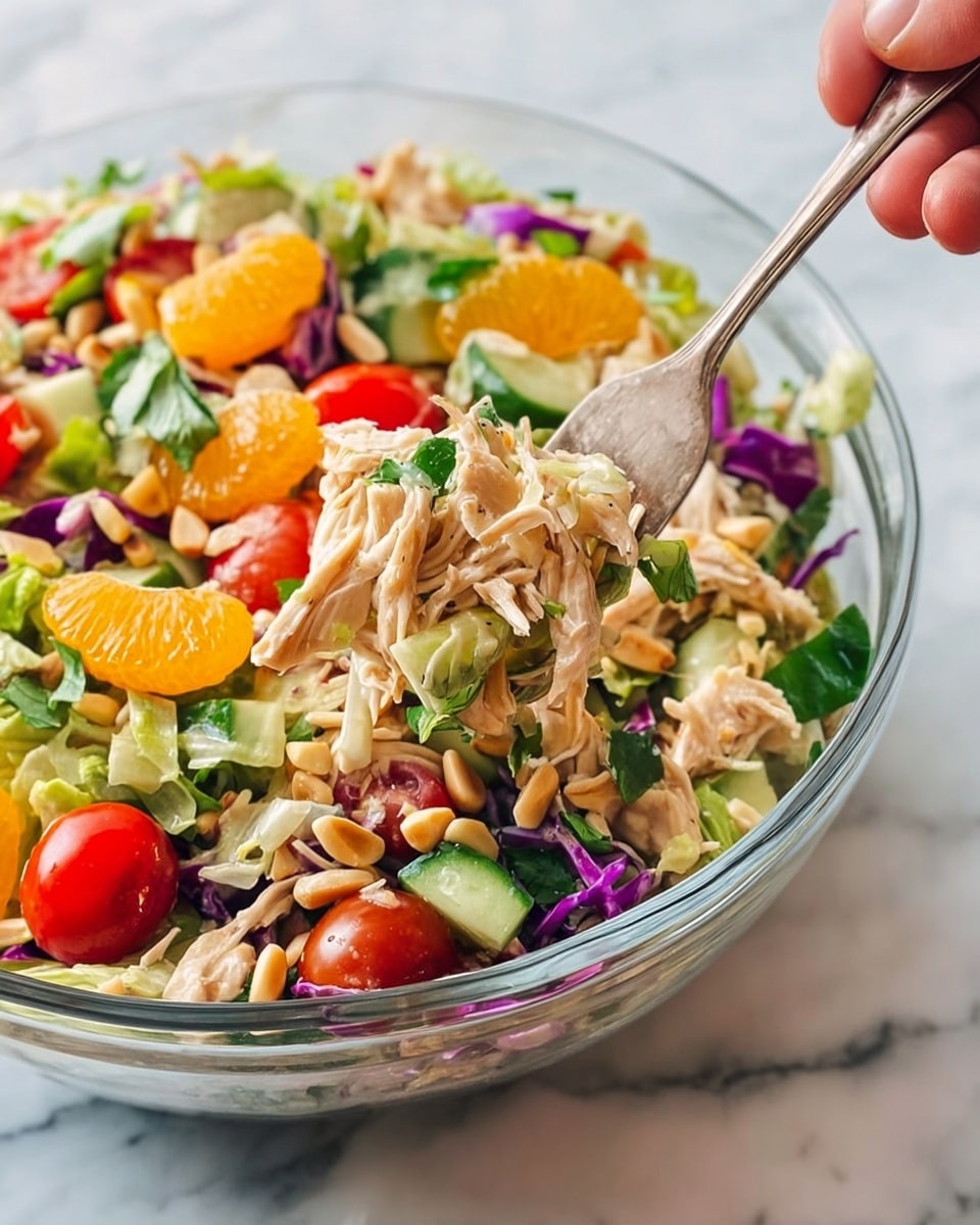 A clear glass bowl filled with a colorful chicken salad sits on a white marbled surface. The salad has several layers: shredded light brown chicken pieces mixed throughout, bright orange mandarin segments scattered on top, halved red cherry tomatoes, chunks of green cucumber, thin slices of purple cabbage, and fresh green leafy herbs. Small pale pine nuts are spread over the whole mix. A silver fork, held by a woman's hand, lifts a forkful of shredded chicken from the bowl, showing the tender texture of the meat mixed with some salad bits. photo taken with an iphone --ar 4:5 --v 7