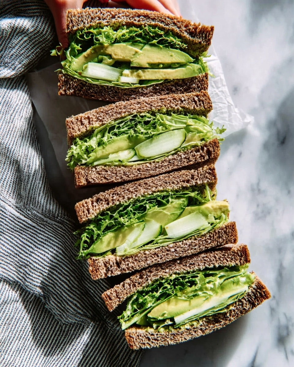A tall sandwich cut in half is stacked on a white plate with green herbs scattered around. The sandwich has three slices of brown bread with light green herb sauce spread on each slice, visible both on the inside and slightly oozing out. Between the bread layers are several layers of fresh green lettuce and leafy greens, thick slices of creamy green avocado, and pieces of grilled white cheese with light brown grill marks. The sandwich is held together on top with a small bamboo skewer twist. In the background, there is a small white bowl with a similar green sauce and a few lemon wedges on the white marbled surface. photo taken with an iphone --ar 4:5 --v 7