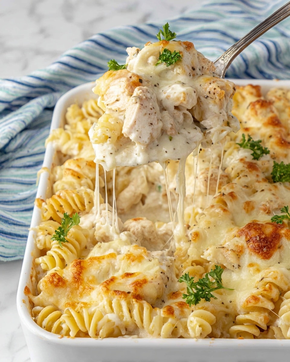 This image shows a white rectangular baking dish filled with creamy baked pasta. The pasta is spiral-shaped and covered in a thick layer of melted cheese that is golden brown in spots. There are pieces of cooked chicken mixed throughout the pasta, coated in a creamy white sauce with black specks, likely pepper. Small green parsley leaves are scattered on top as garnish. A metal spoon is lifting a portion of the pasta, showing melted cheese stretching from the dish. The dish sits on a white marbled surface with a blue and white striped cloth in the background. Photo taken with an iphone --ar 4:5 --v 7