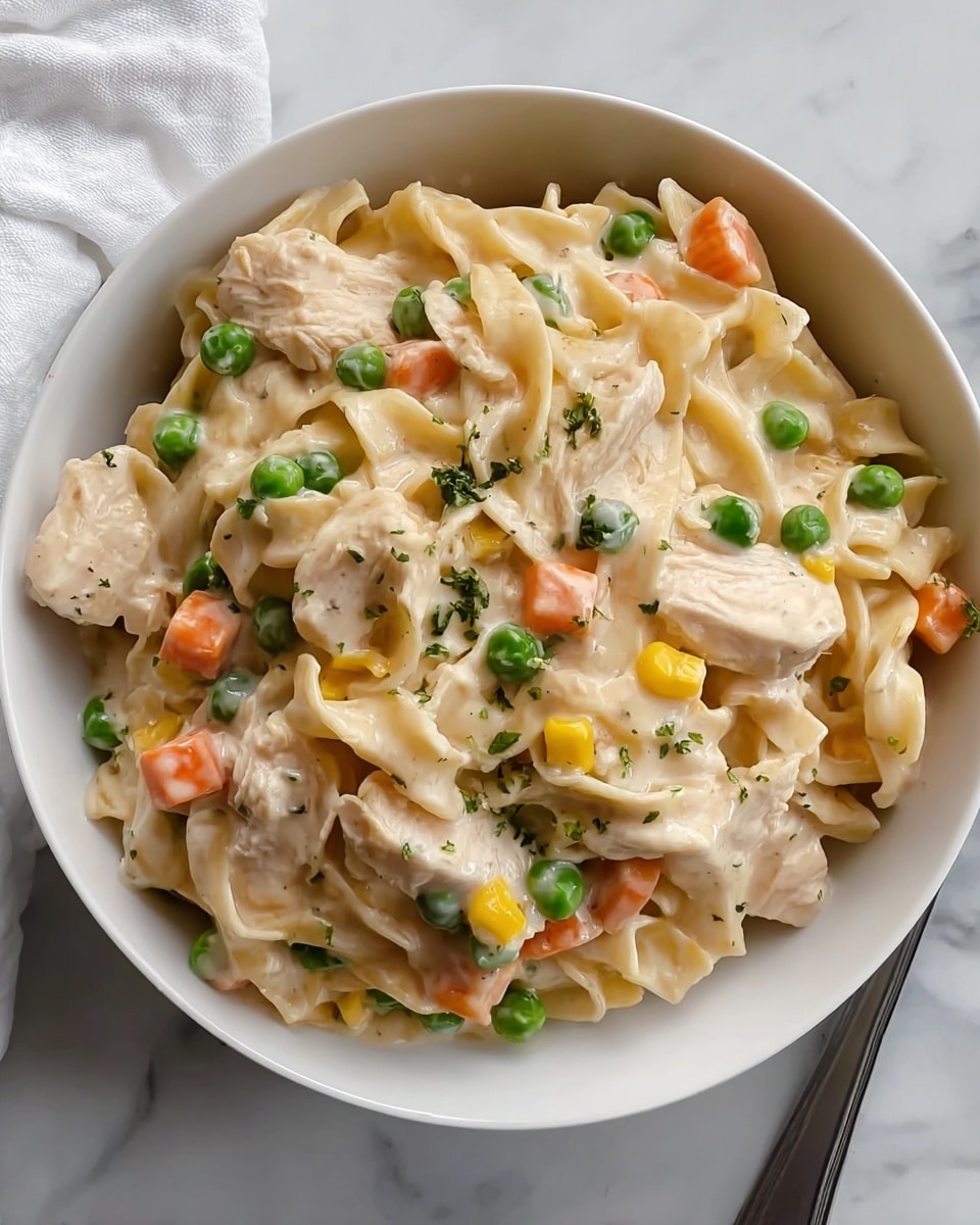 A white bowl filled with creamy chicken and noodle casserole featuring visible layers of small pasta pieces mixed with chunks of light brown cooked chicken, bright green peas, orange carrot cubes, and yellow corn kernels all coated in a smooth, thick, light cream sauce with green herb specks, sitting on a white marbled surface with a white cloth nearby and a stack of white plates and a white vase with white flowers blurred in the background, photo taken with an iphone --ar 4:5 --v 7
