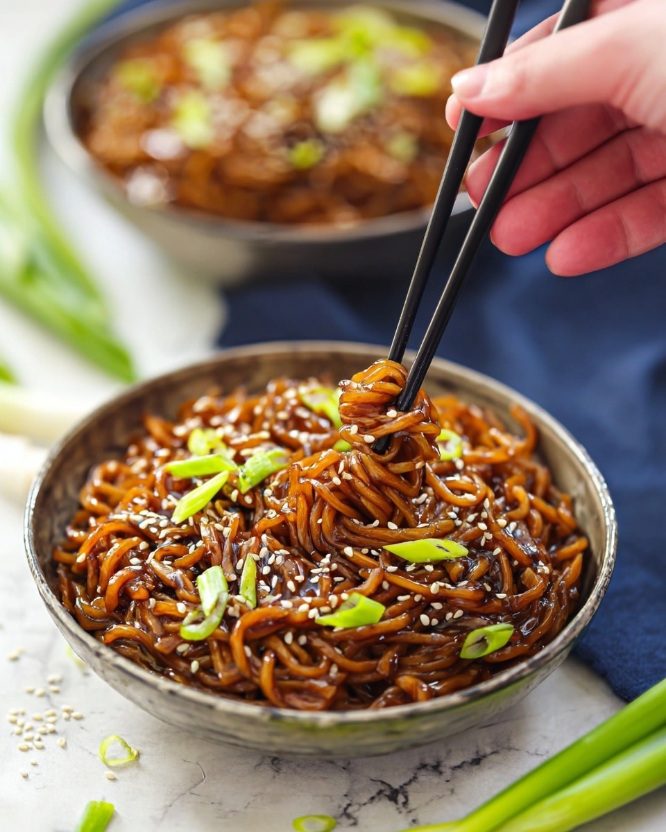 A bowl filled with a large serving of cooked noodles that have a light brown sauce coating, giving them a shiny texture, with small bits of caramelized onions or garlic mixed in. The noodles are topped with sliced bright green scallions and sprinkled with small white sesame seeds. The bowl is a speckled gray ceramic, sitting on a wooden surface beside a pair of wooden chopsticks. In the background, a small bowl with dark red sauce and a silver spoon can be seen, along with a small dish of sesame seeds. photo taken with an iphone --ar 4:5 --v 7
