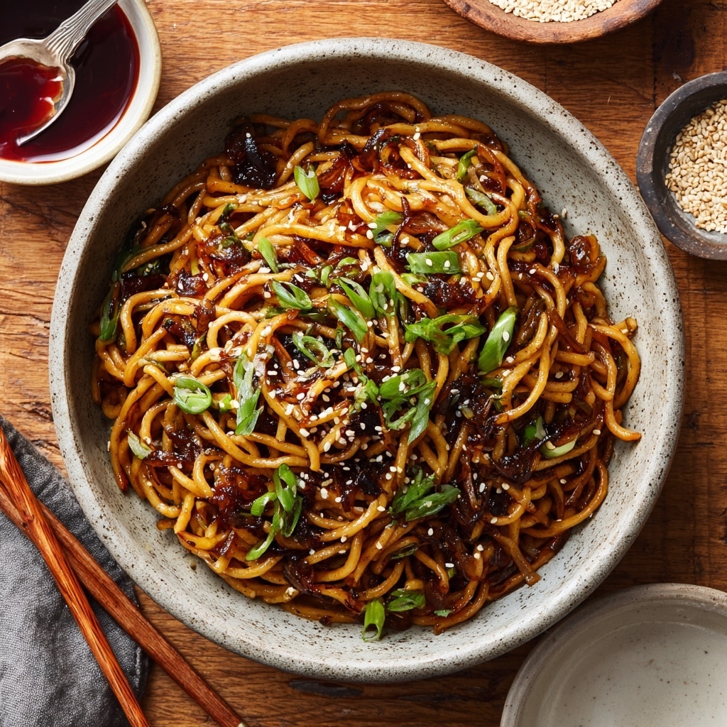 A close-up image shows a silver bowl filled with glossy, dark brown noodles coated in thick sauce, garnished with scattered white sesame seeds and small, bright green chopped scallions. A woman's hand holding black chopsticks picks up a tangle of noodles, emphasizing the texture and shine of the dish. In the background, there is a blurred second bowl with similar noodles and scallions. Green scallion stalks and scattered sesame seeds are visible on the white marbled surface beneath the bowls. The scene has a fresh and appetizing look. photo taken with an iphone --ar 4:5 --v 7