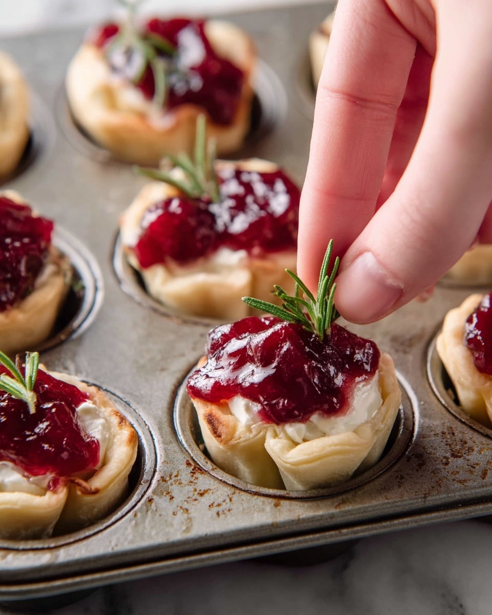 A close-up shot of six mini tartlets in a metal muffin pan, each with three visible layers: the bottom layer is a light golden dough base folded at the edges inside each cup, topped with a thick white creamy cheese layer, and finished with a glossy, chunky deep red cranberry sauce on top. A woman's hand is placing a small green rosemary sprig into one of the tartlets, adding a fresh green touch. The pan shows some signs of use with slight stains, all set against a white marbled texture. Photo taken with an iphone --ar 4:5 --v 7