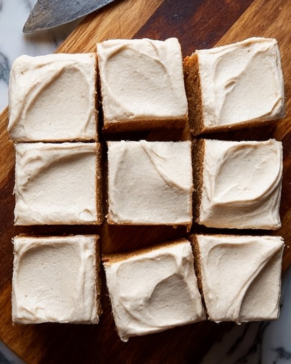 A square cake cut into nine pieces sits on a wooden surface replaced by a white marbled texture. The cake has one thick layer of light brown base covered with a smooth, creamy, off-white frosting spread evenly across the top with soft swoops and slight ridges. Each piece reveals the contrast between the darker cake and the lighter frosting layer. The edges of the frosting are slightly uneven, showing a gentle, homemade texture. Photo taken with an iphone --ar 4:5 --v 7