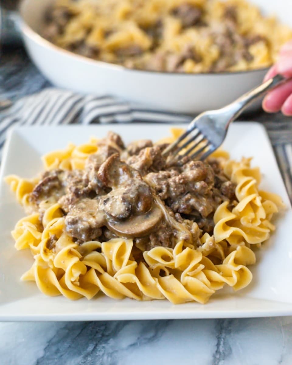 The image shows a close-up of a white square plate filled with a creamy beef stroganoff dish. The dish has two main layers: the base layer is made of wavy yellow egg noodles with a soft texture, and the top layer contains pieces of cooked ground beef mixed with sliced brown mushrooms covered in a light creamy sauce. The background shows a white bowl with more of the same dish, slightly out of focus, all set on a white marbled surface. A woman's hand is holding a fork near the edge of the bowl. Photo taken with an iphone --ar 4:5 --v 7
