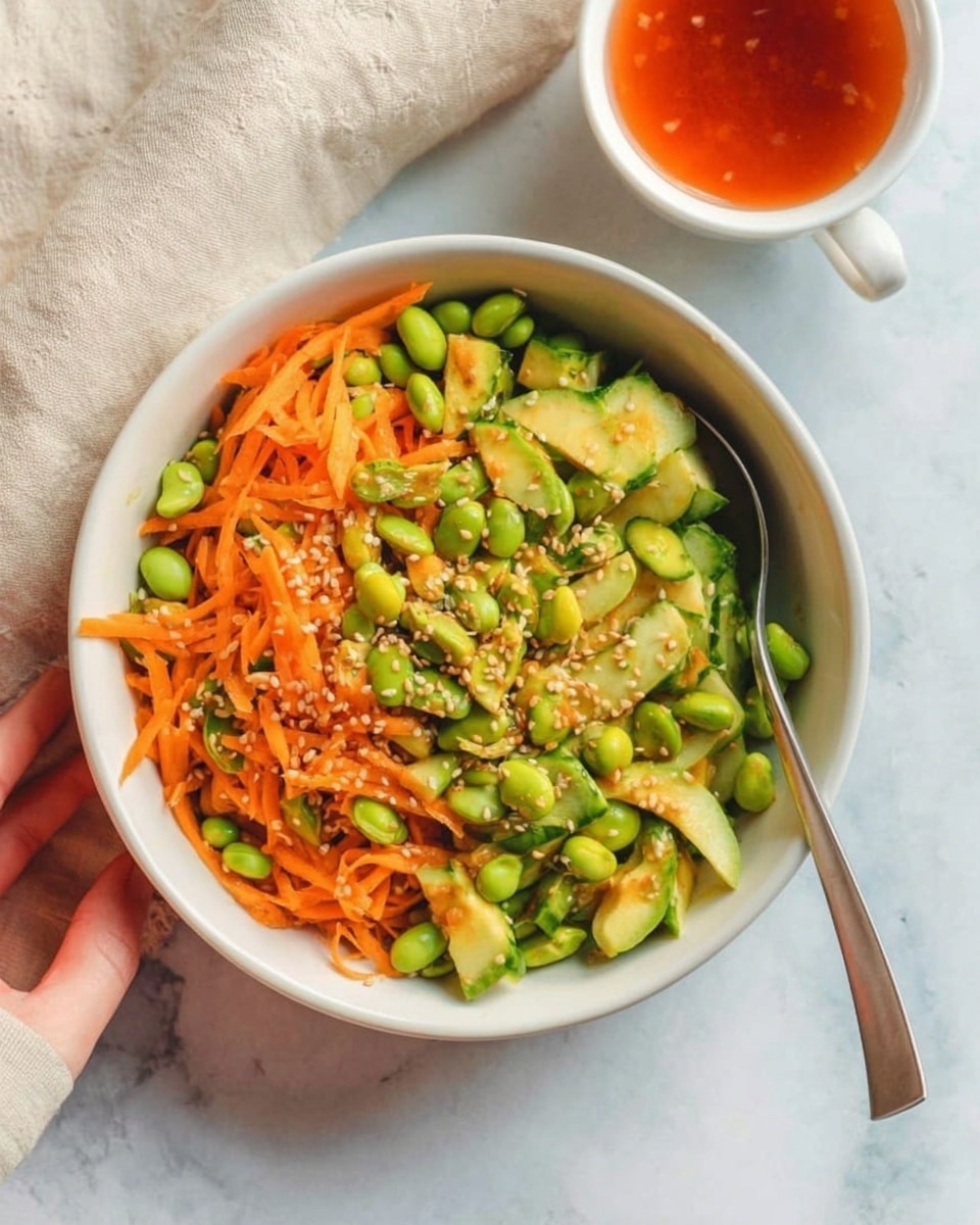 A white bowl filled with a colorful salad featuring three main layers: the bottom layer has bright green edamame beans scattered throughout, the middle layer consists of thinly sliced cucumber pieces in fresh green hues with slight shine, and the top layer is made of thin, vibrant orange carrot sticks mixed evenly across the bowl; small white sesame seeds are sprinkled on top for texture and visual contrast. To the right of the bowl, a silver spoon rests inside, and next to it is a white cup with reddish-orange dressing inside, sitting on a white marbled surface. A woman's hand is holding the edge of the bowl on the left side gently. The scene has a natural light setting, clean and fresh with a white marbled background. photo taken with an iphone --ar 4:5 --v 7