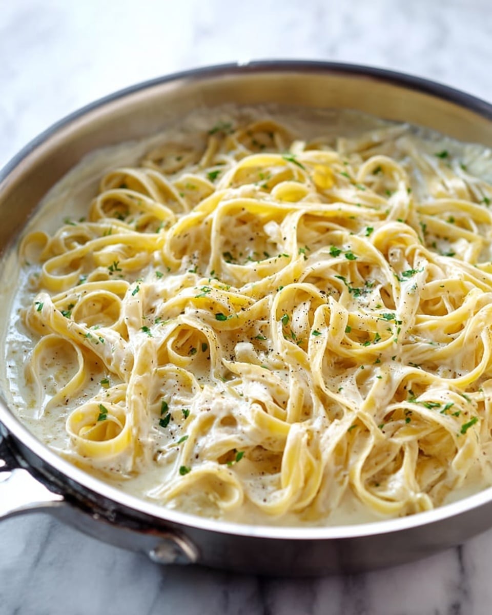 A close-up view of a silver pan filled with creamy white sauce coating long, flat, pale-yellow fettuccine noodles, lightly sprinkled with small green parsley pieces and black pepper. The sauce looks smooth and rich, pooling a bit at the bottom while the noodles twist and curl naturally inside the pan. The pan sits on a white marbled surface with soft natural light highlighting the glossy texture of the sauce and noodles. photo taken with an iphone --ar 4:5 --v 7