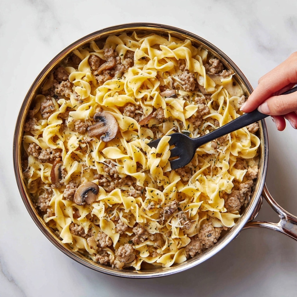 A close-up image of a pan filled with cooked egg noodles mixed with browned ground meat and sliced mushrooms. The noodles are twisted and layered unevenly, showing a soft texture and a light yellow color. The brown meat pieces and the darker brown mushrooms are spread throughout the noodles, creating a mix of colors and textures. The pan has a polished silver rim and is placed on a white marbled surface. A woman's hand is using a black fork to lift some noodles from the pan. Photo taken with an iphone --ar 4:5 --v 7