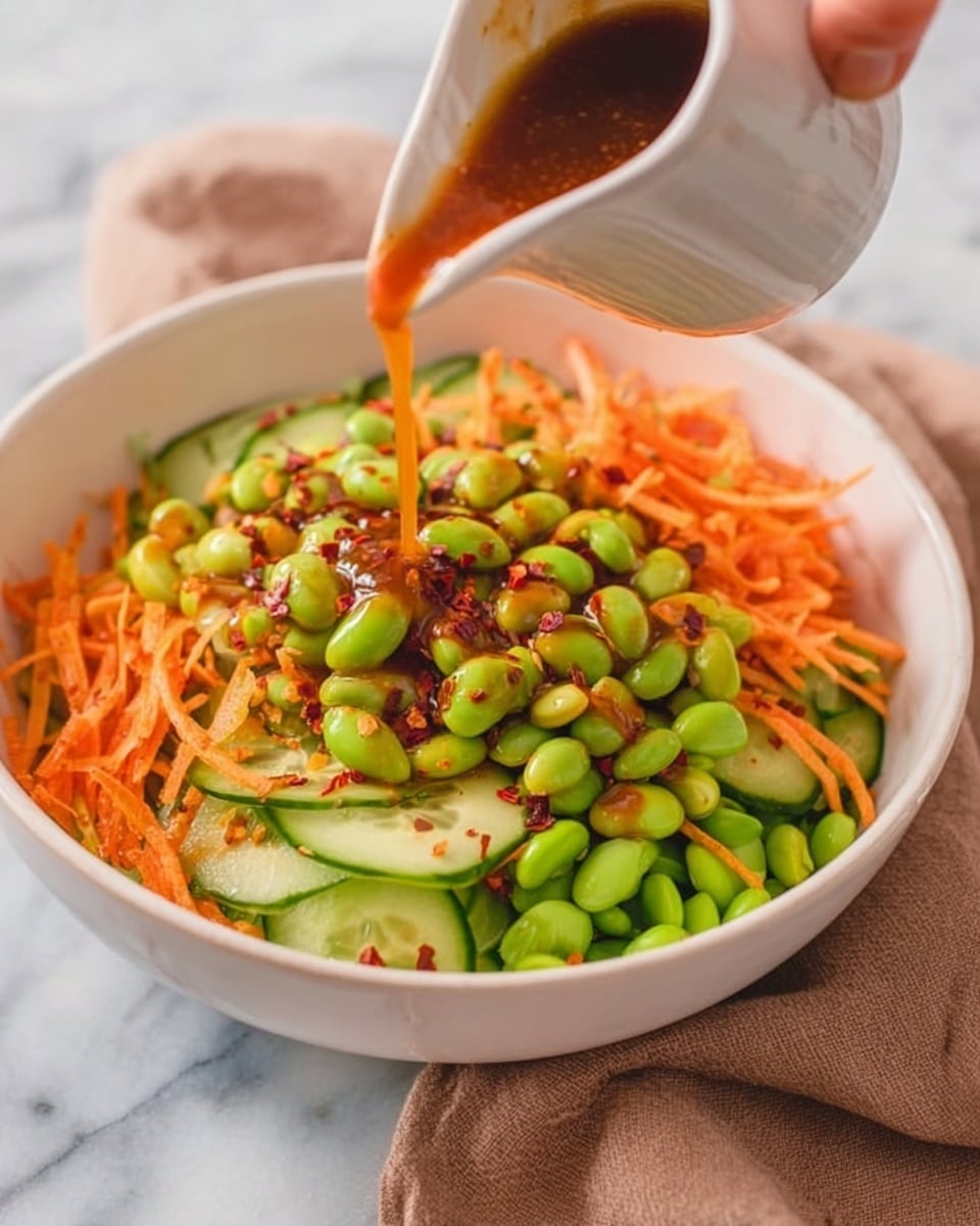 A white bowl filled with a fresh salad, showing several layers: the bottom layer has bright green edamame beans, followed by light green cucumber slices with dark green edges, and topped with thin, shredded orange carrots. Red chili flakes are scattered over the salad. A woman's hand is pouring a reddish-brown dressing from a small white pitcher over the salad. The bowl sits on a white marbled surface, with a soft brown cloth nearby. Photo taken with an iphone --ar 4:5 --v 7