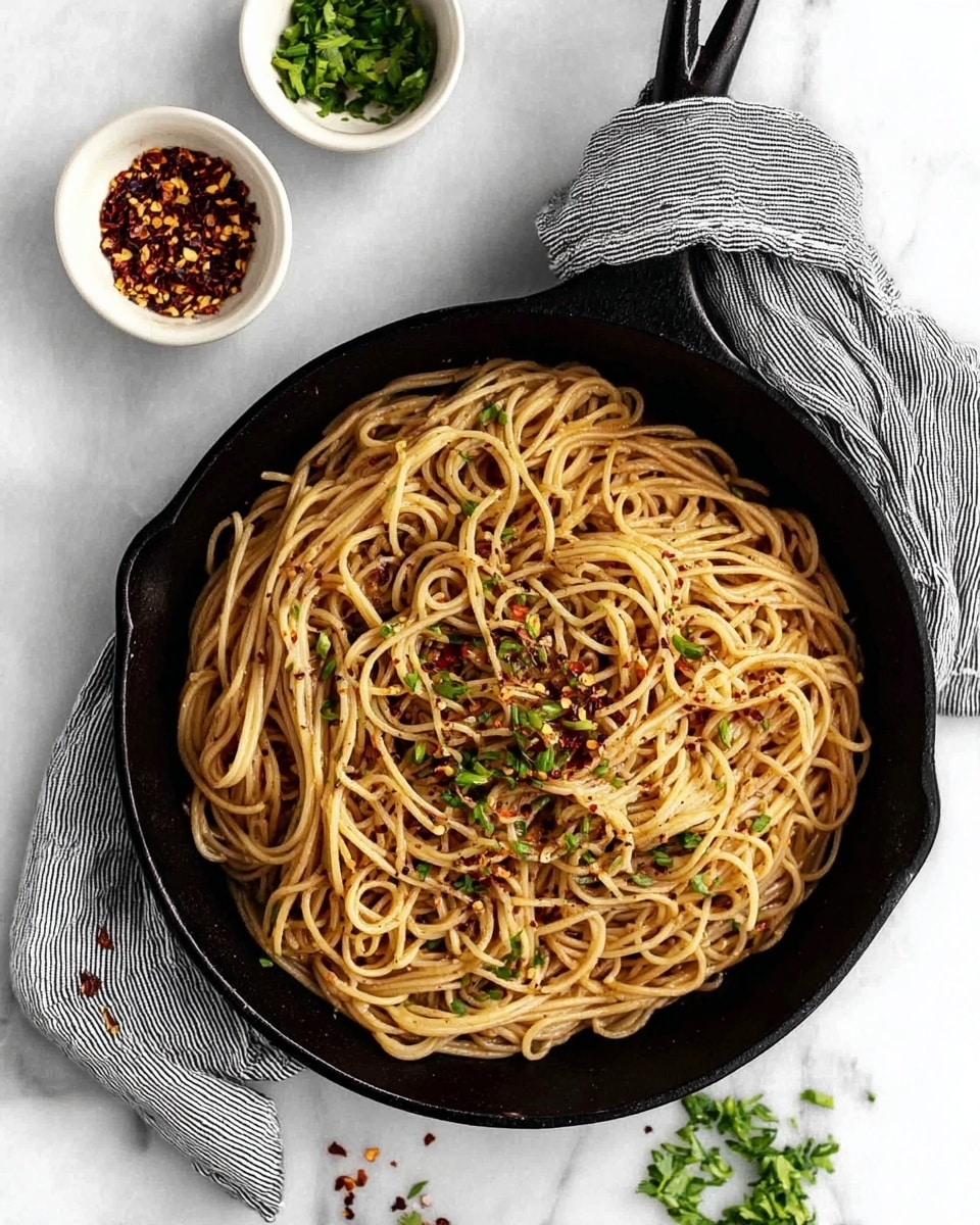 A black cast iron skillet filled with light brown cooked noodles that have a slightly shiny texture, topped with small green chopped herbs and red chili flakes sprinkled across. The skillet handle is wrapped in a gray and white striped cloth. Nearby, two small white bowls sit on a white marbled surface; one bowl contains more red chili flakes and the other has chopped green herbs. Some herbs are scattered on the white marbled surface around the skillet. Photo taken with an iphone --ar 4:5 --v 7