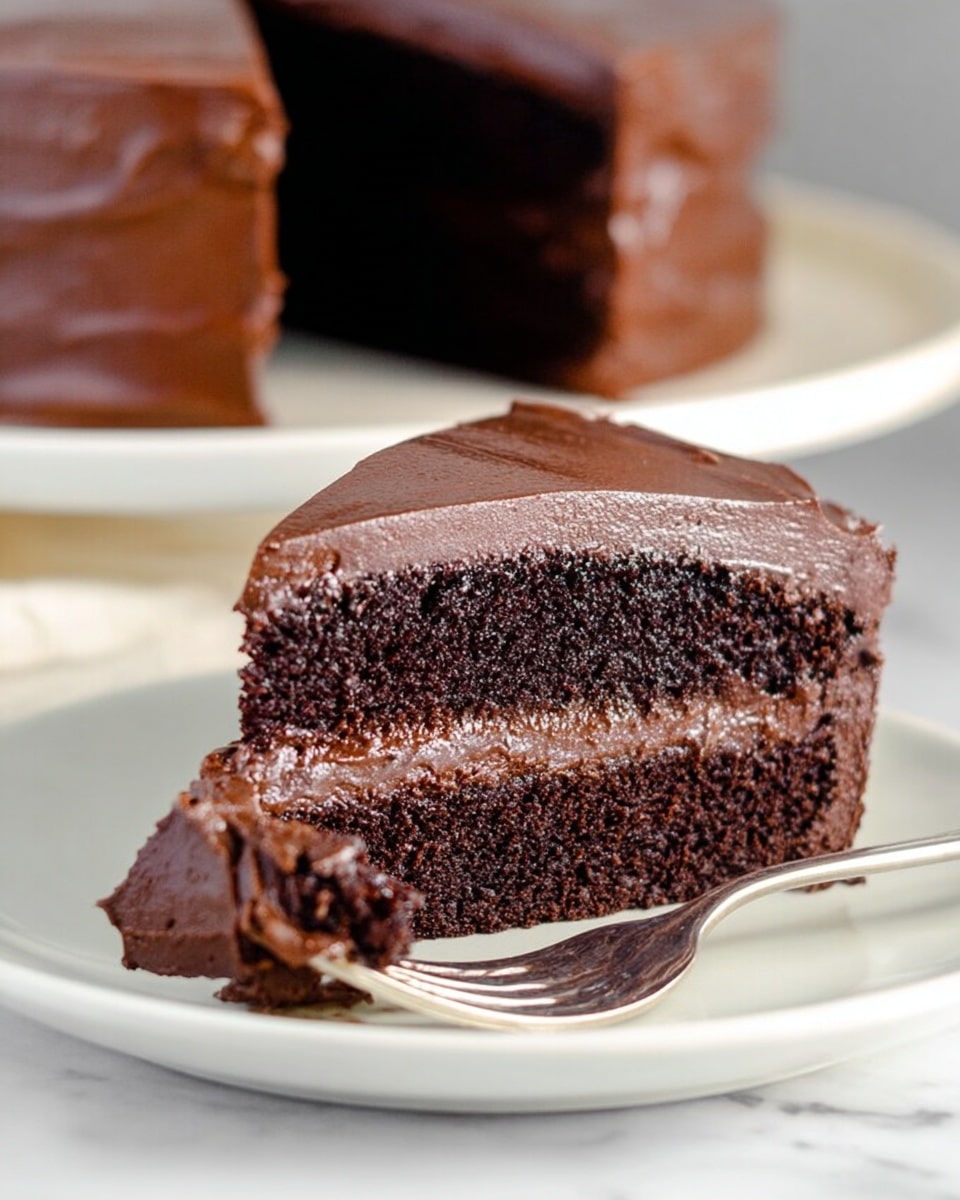 A close-up view of a thick slice of chocolate cake with three layers, dark brown and moist with a slightly crumbly texture. Between each cake layer is a smooth, shiny middle layer of lighter chocolate frosting. The outside of the slice is covered with a rich, smooth chocolate frosting that looks creamy and glossy. The slice is placed on a simple white plate atop a white marbled surface. In the background, a whole chocolate cake with the same smooth frosting is slightly out of focus, giving depth to the image. Photo taken with an iphone --ar 4:5 --v 7