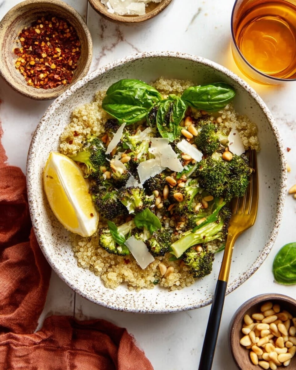A white speckled bowl holds a dish with three main layers: the bottom layer is light beige quinoa with a fluffy texture, the middle layer consists of bright green roasted broccoli florets with slightly browned edges scattered evenly, and the top layer is sprinkled with golden pine nuts and pale, thin shavings of cheese, garnished with fresh green basil leaves. A fresh lemon wedge rests on the side inside the bowl. A gold and black fork is placed on the right edge of the bowl. The bowl sits on a white marbled surface surrounded by small bowls containing pine nuts, fresh basil leaves, and red pepper flakes, with a glass of amber liquid and a rust-colored cloth napkin nearby. Photo taken with an iphone --ar 4:5 --v 7