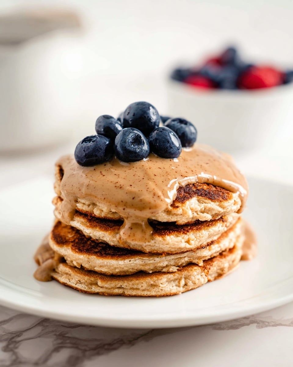 A stack of four thick, fluffy pancakes with a golden-brown color sits at the center of a white plate. The pancakes have a soft, slightly porous texture with a darker, crispy edge. On top, creamy brown sauce drips slightly over the sides, smooth and glossy with a speckled look. A cluster of five plump, deep blue blueberries rests on the sauce, adding a fresh, round contrast. The plate is on a white marbled background, with a blurred white bowl filled with berries in the background. Photo taken with an iphone --ar 4:5 --v 7