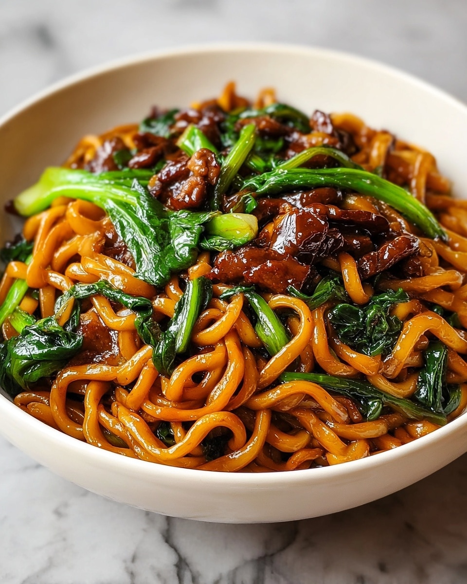 The image shows a close-up of a pile of thick yellowish-brown noodles mixed with dark brown pieces of meat and bright green leafy vegetables, all cooked together and glistening with a shiny sauce. The noodles are thick and slightly curled, layered throughout the dish, with the green vegetables and meat pieces spread evenly among them. The food is served in a large dark metal wok with a slightly rough texture. photo taken with an iphone --ar 4:5 --v 7