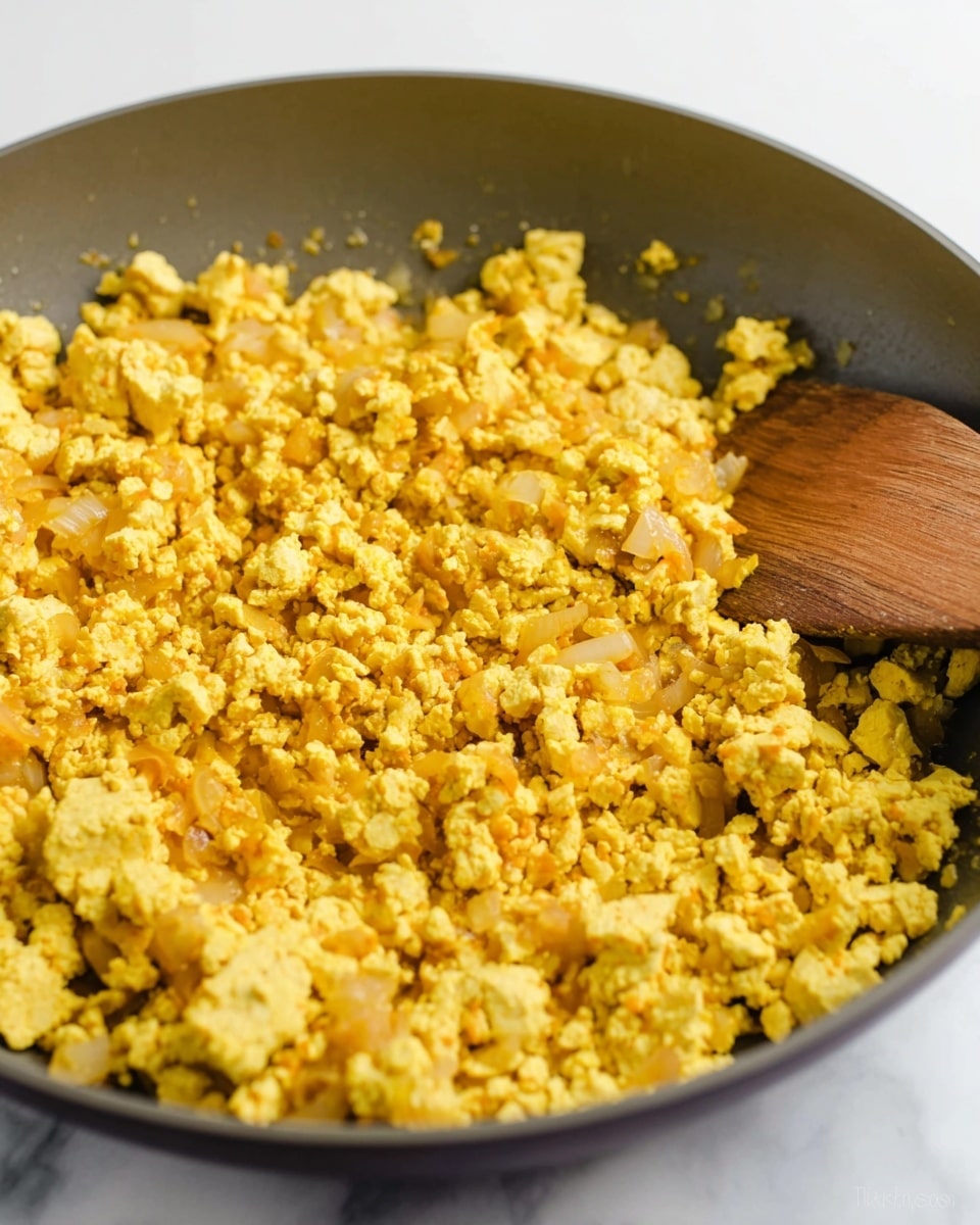 A close-up view of a pan filled with one main layer of soft, scrambled tofu mixed with small pieces of sautéed onions. The tofu is golden yellow with slightly browned edges, showing a crumbly texture. The onions are translucent with a light golden tint, blending evenly throughout the tofu. A wooden spatula rests on the right edge of the pan, which is a dark gray color, sitting on a white marbled surface. The image captures the warm, cooked texture of the tofu scramble up close with bright lighting. photo taken with an iphone --ar 4:5 --v 7