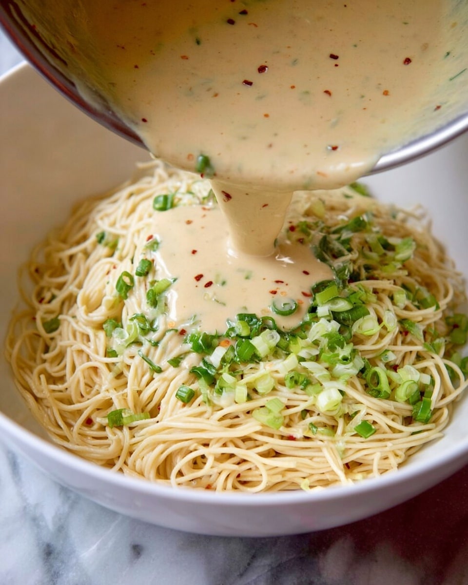 A white bowl filled with creamy, light beige noodles mixed with small green herbs and dark black sesame seeds, topped with fresh bright green cilantro leaves scattered around. The noodles look thick and slightly coated with sauce, giving a smooth texture. Black and beige chopsticks hold a small bundle of noodles on the left side of the bowl. A small white sauce bowl with a light brown creamy sauce is in the upper left corner on a white marbled surface. Photo taken with an iphone --ar 4:5 --v 7