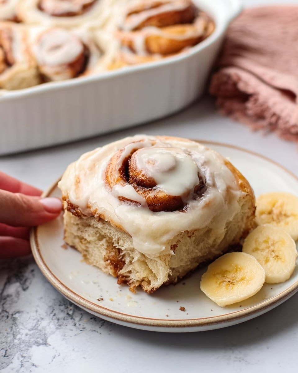 The image shows a close-up of a single cinnamon roll on a white plate with two slices of banana on the side. The cinnamon roll has three visible layers: a thick, soft-looking dough base that is light brown with swirls of darker cinnamon, a gooey cinnamon filling in between the layers, and a smooth, creamy white icing on top that drips slightly down the sides. The plate is set on a white marbled surface, and a woman's hand is gently touching the cinnamon roll from the left side. In the background, there is a white baking dish filled with more cinnamon rolls, slightly blurred. photo taken with an iphone --ar 4:5 --v 7