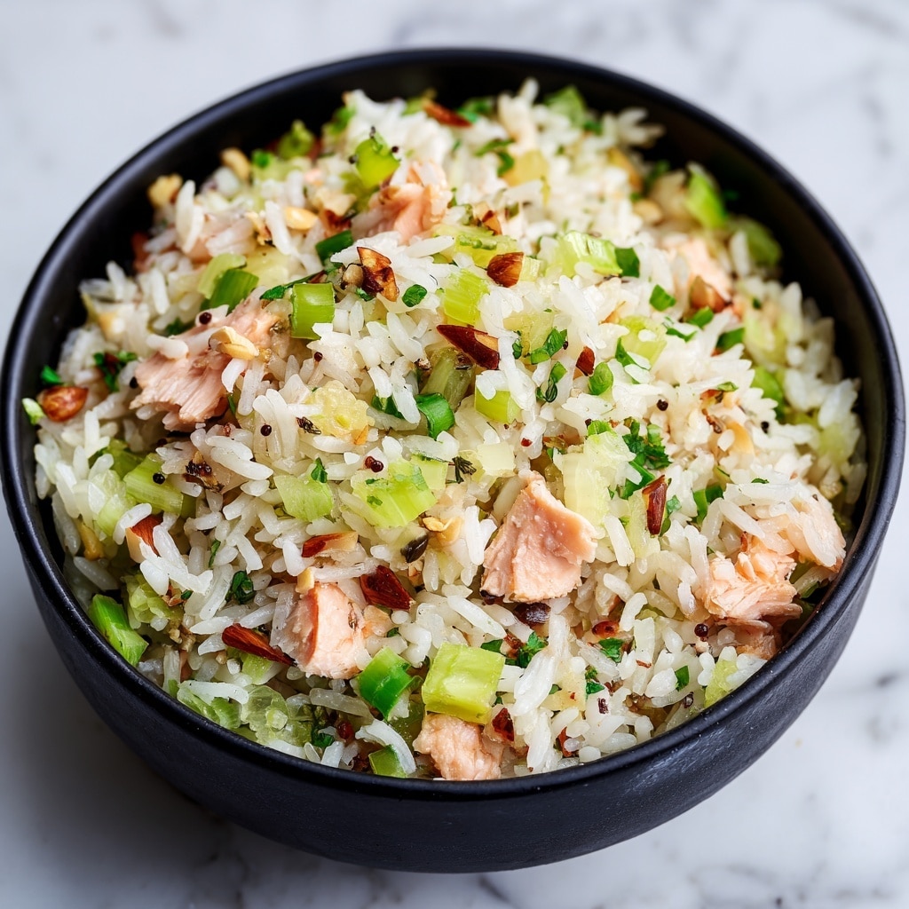 A clear glass bowl filled with a mixed rice salad sits on a white marbled surface. The dish has several layers of white rice grains mixed evenly with light green chopped celery, bright green sliced scallions, and small pieces of light pink cooked salmon. Scattered throughout are small brown toasted almonds adding texture, and tiny black seeds dot the mix, giving it a speckled look. The ingredients appear fresh and finely chopped, creating a colorful and light-textured meal. photo taken with an iphone --ar 4:5 --v 7