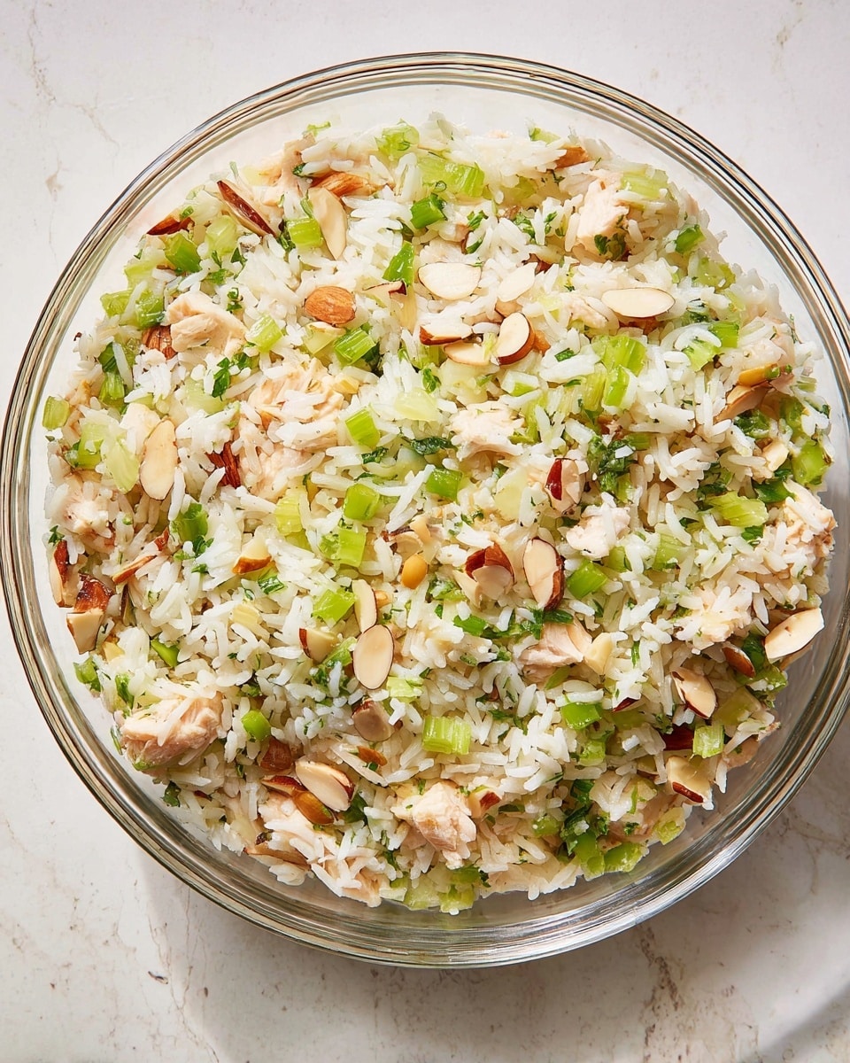 A clear glass bowl contains a mixed rice dish with three main layers visible: the base layer of white rice, the middle layer scattered with light pink pieces of cooked chicken, and the top layer mixed with chopped green celery, green onions, and whole almonds, all evenly distributed throughout. The textures range from the fluffy rice grains, tender chicken chunks, crisp celery, and crunchy almonds, giving a fresh and colorful look. The bowl sits on a white marbled textured surface. Photo taken with an iphone --ar 4:5 --v 7