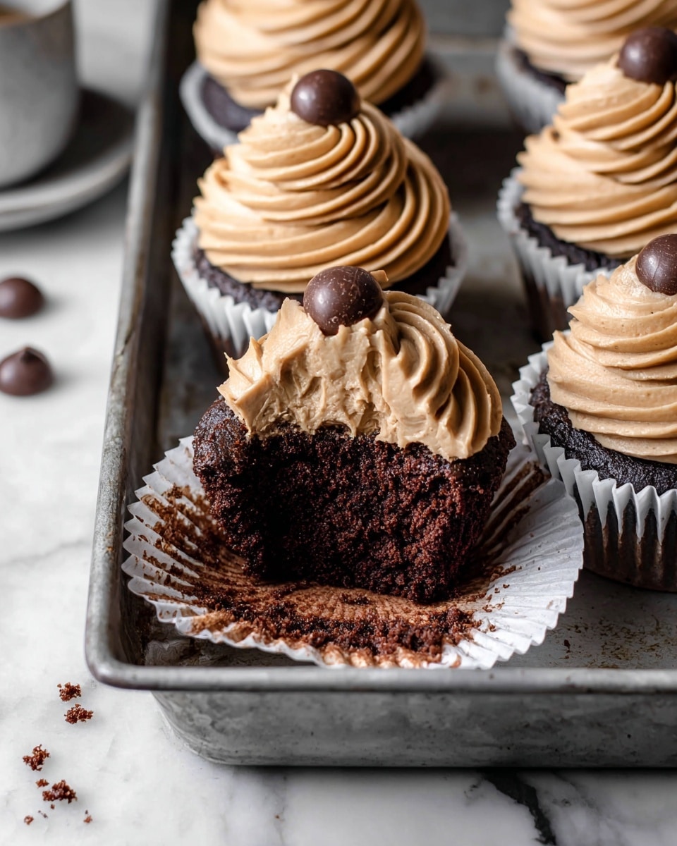 The image shows chocolate cupcakes with two layers: a dark, moist chocolate cake base topped with a smooth light brown frosting swirled in a circular pattern. Each cupcake is wrapped in a white paper liner, and some have a single dark chocolate chip placed on top of the frosting swirl. One cupcake is open, revealing the dense chocolate layer beneath the creamy frosting. The cupcakes sit in a metal tray on a white marbled surface, with a few chocolate crumbs scattered around. photo taken with an iphone --ar 4:5 --v 7