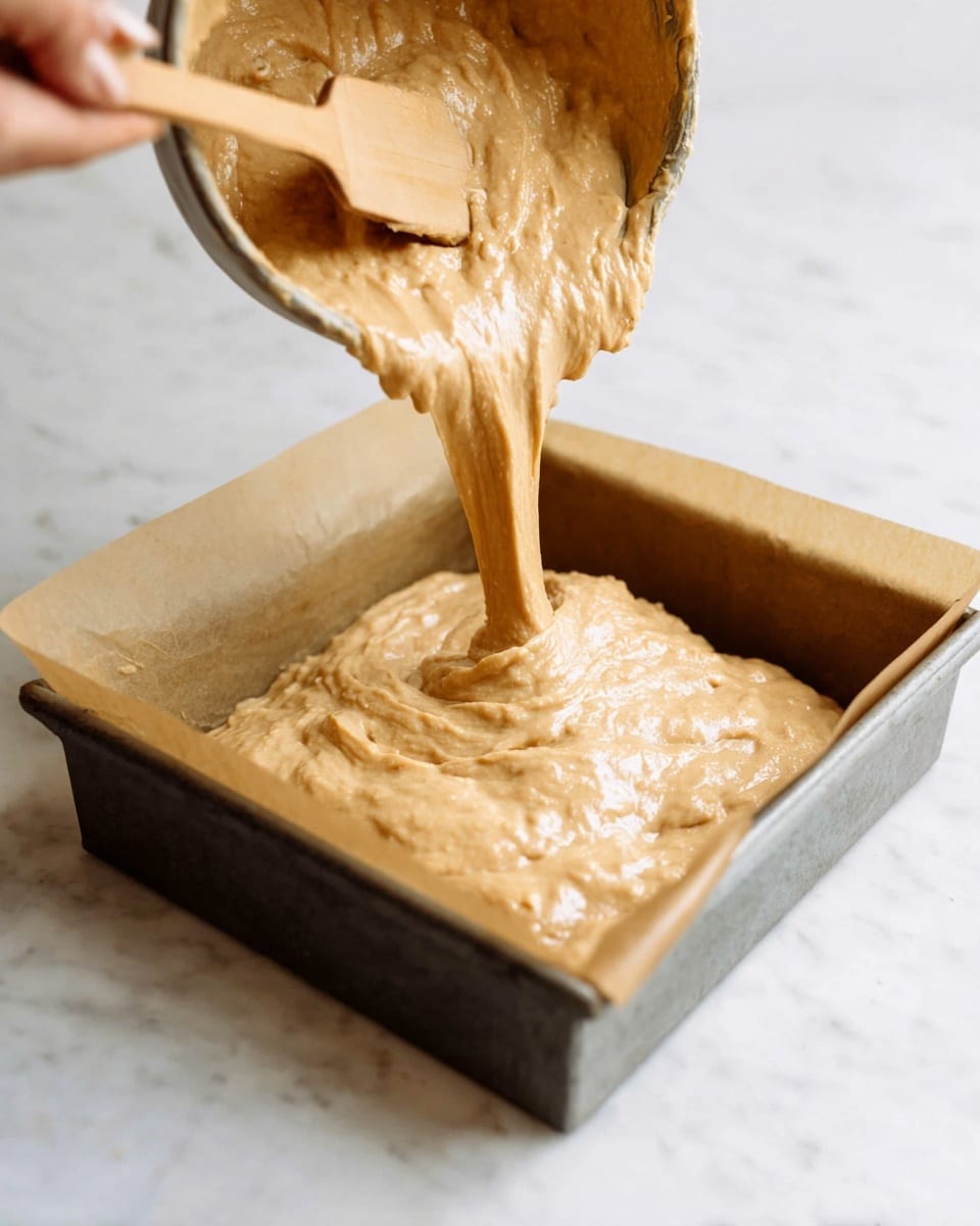 A close-up view of a square piece of cake with two layers, placed in front of similar cake pieces on a sheet of parchment paper. The bottom layer is a light brown, moist cake with visible tiny dark specks inside, showing a soft and crumbly texture. The top layer is smooth white frosting spread thickly and topped with small chunks of light brown walnuts. Some walnut pieces and crumbs are scattered on the white marbled surface around the cake. In the foreground, a spoon holds a small amount of white frosting with walnut bits. The background is softly blurred with a hint of a small container tilted on its side. Photo taken with an iphone --ar 4:5 --v 7